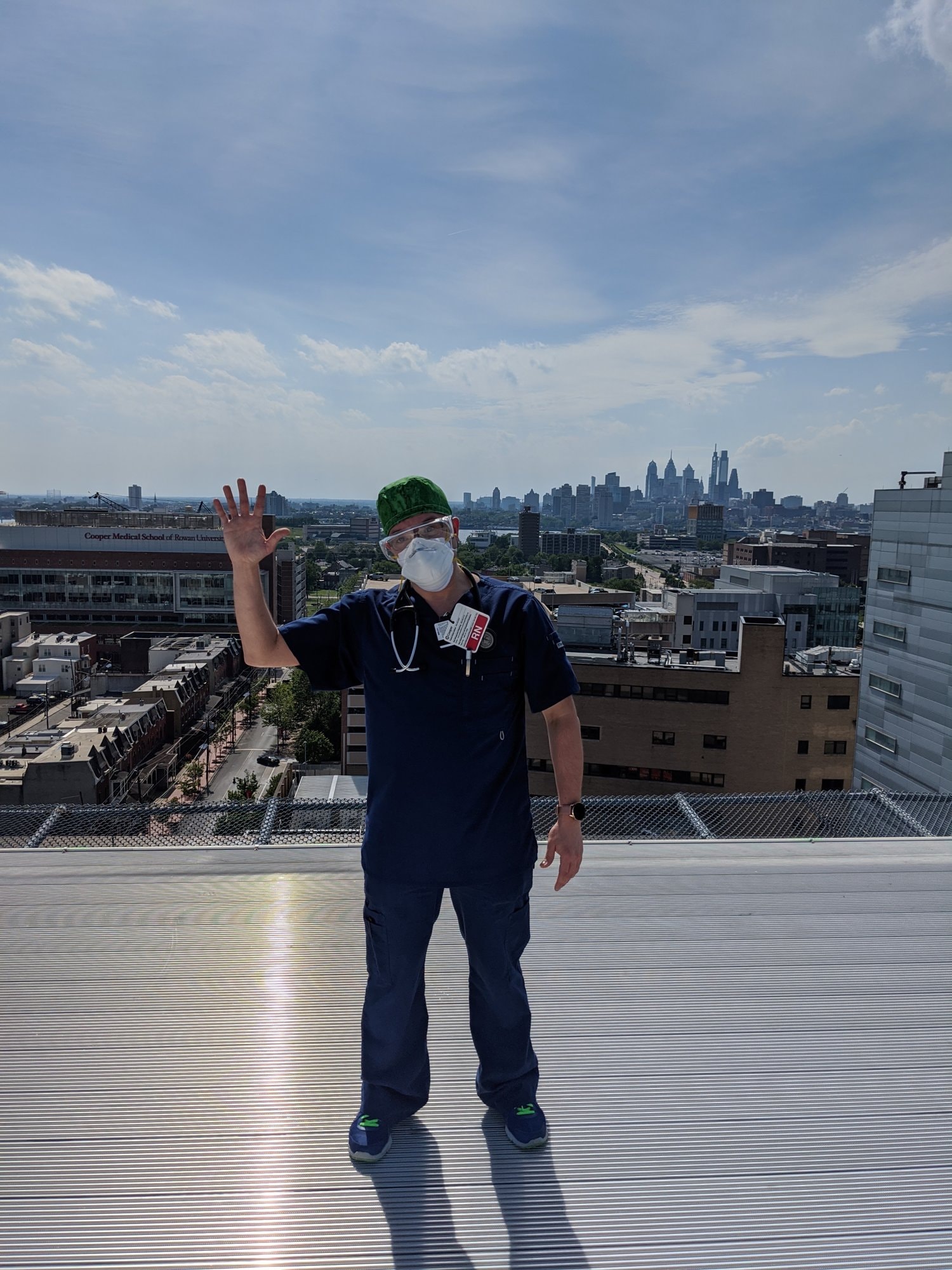 Travis in scrubs, an N95 mask and goggles, waving from a hospital rooftop with the Philadelphia skyline behind him